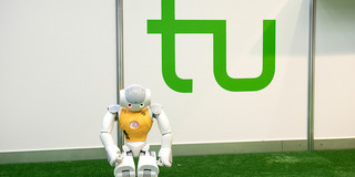 Fußballroboter vor TU-Logo Ein Roboter mit gelbem Trikot steht an der Seitenlinie eines Fußballfelds, hinter ihm ist das grüne Logo der TU Dortmund.